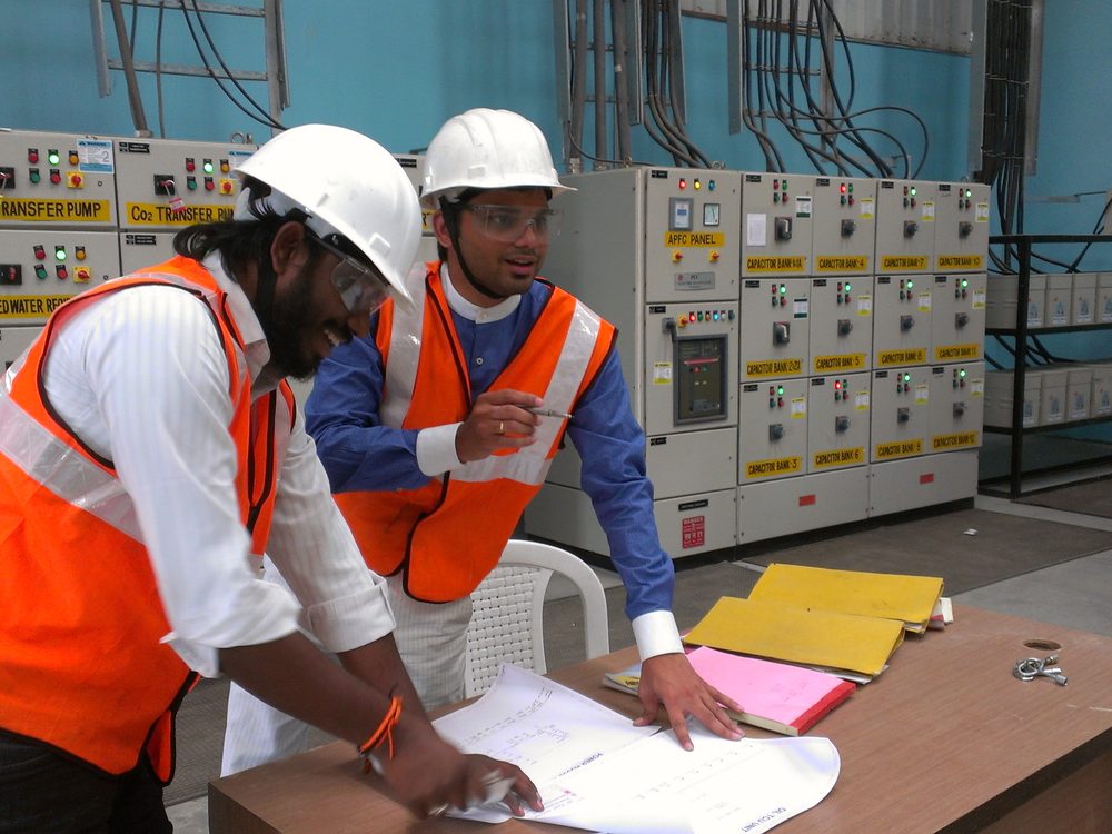 Two engineers reviewing a single line diagram against labelled switchgear during a plant walkdown